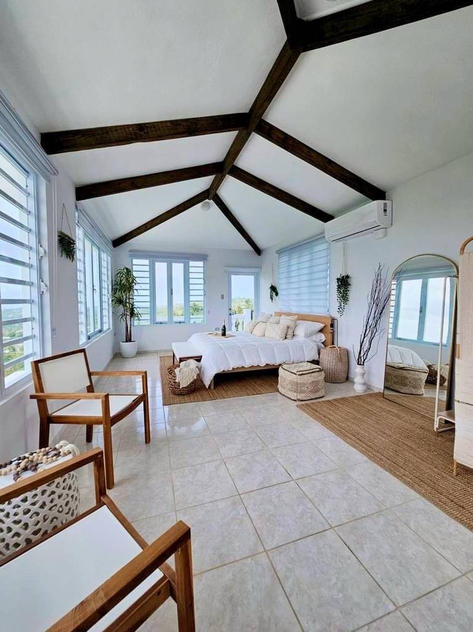 A large corner bedroom under a vaulted ceiling crossed with dark timber beams, with windows on three sides