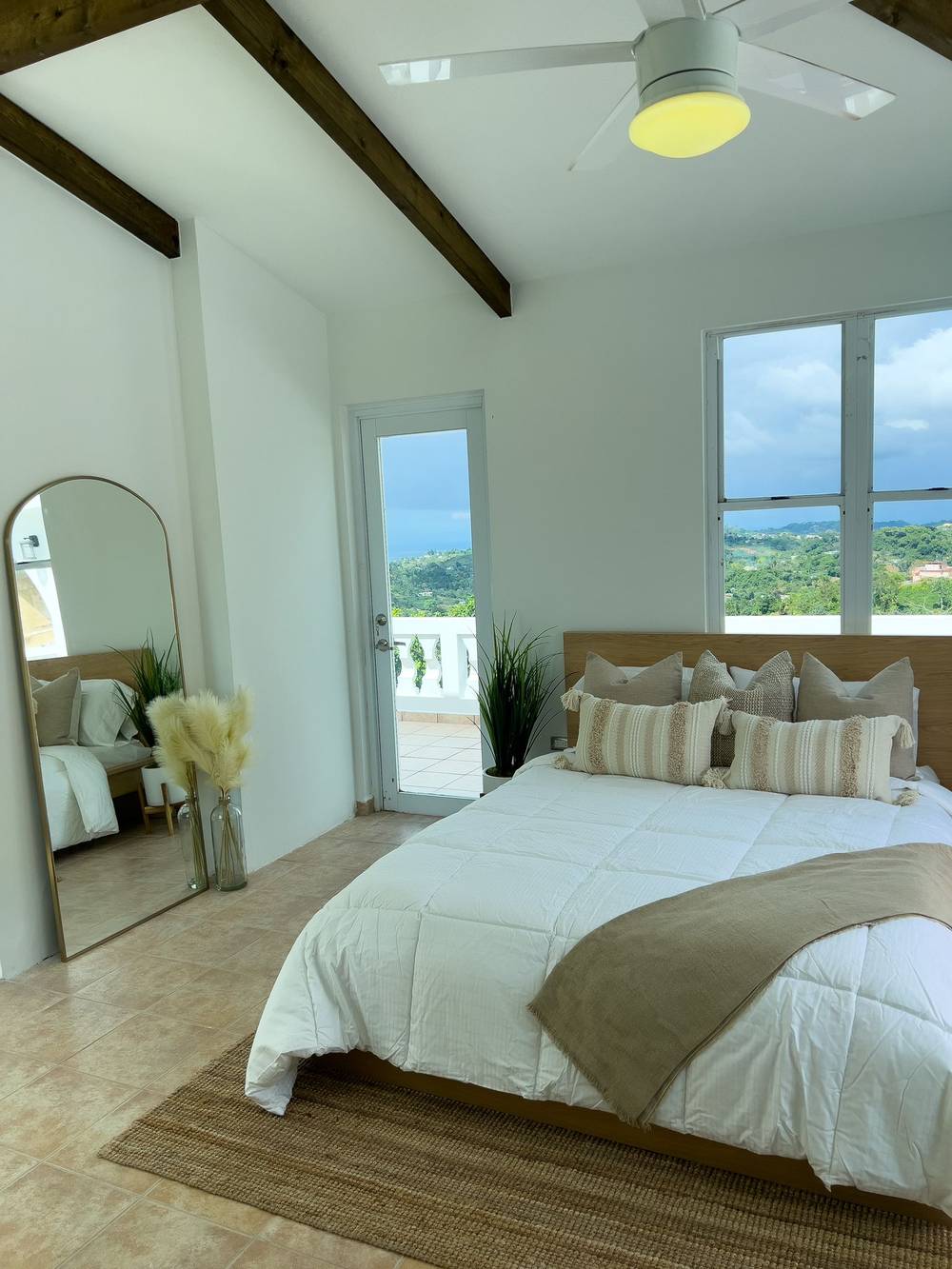 A bright bedroom with exposed beams, a tall arched mirror and a glass door opening onto the balcony
